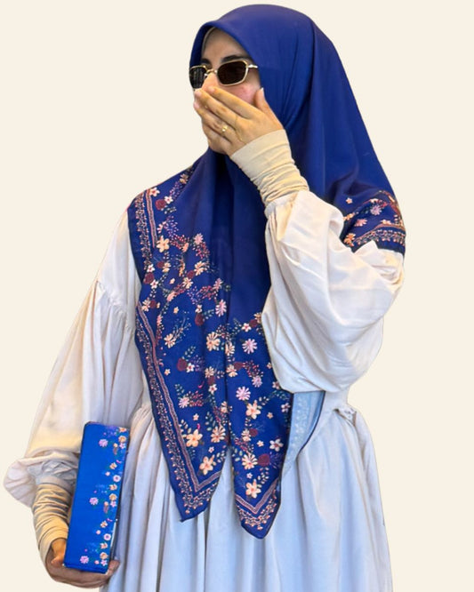 A woman wearing a royal blue hijab with white floral embroidery and a white long-sleeve dress, holding a matching blue clutch against a plain beige background.