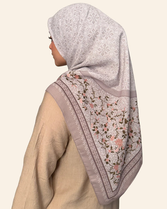 A woman shown in profile wearing a light grayish-lavender hijab with a floral print and embroidered border, paired with a beige top against a plain cream background.