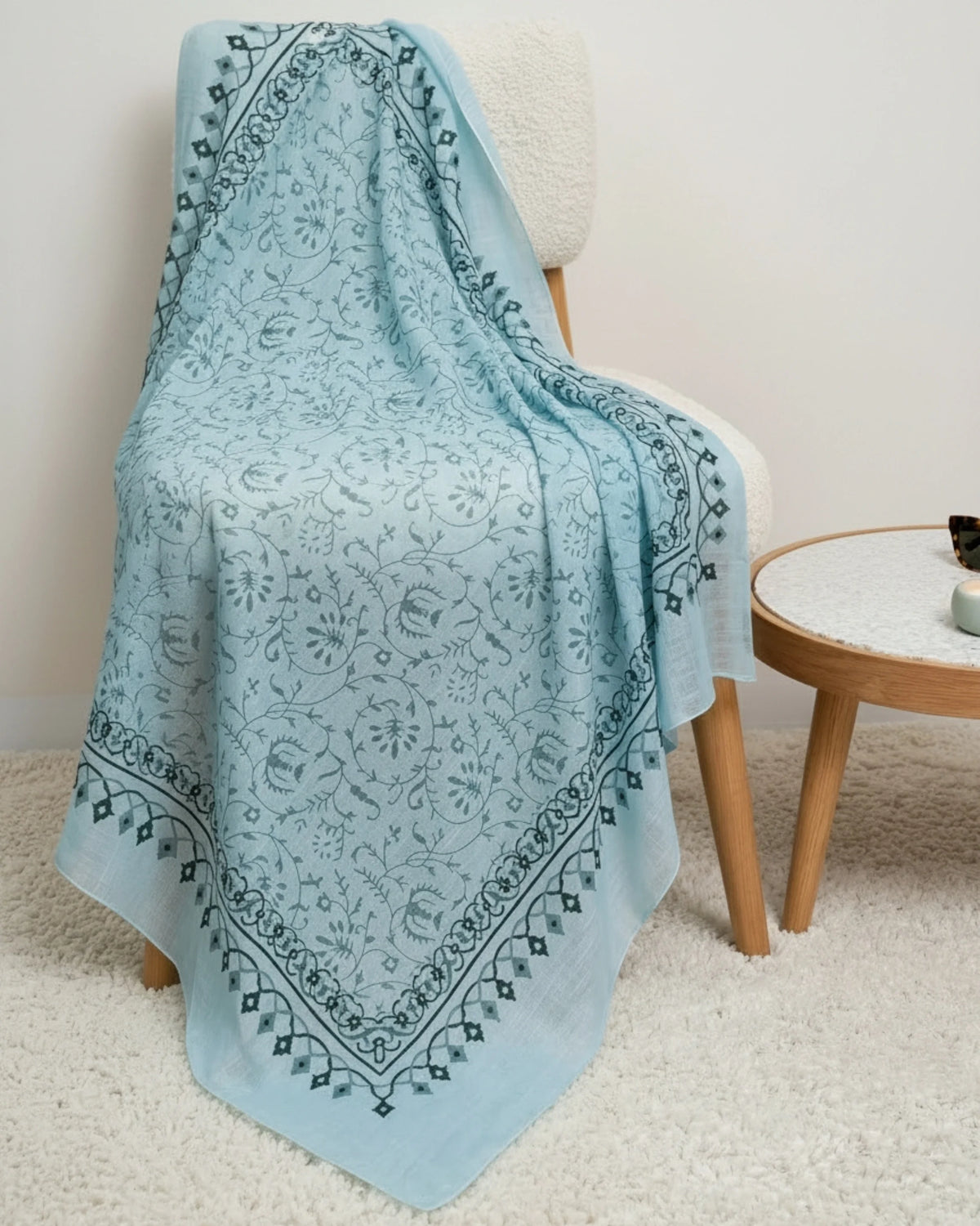 A light blue bandana throw blanket with a dark blue botanical border and central floral pattern, draped over a cream-colored armchair in a cozy room setting next to a small wooden side table.