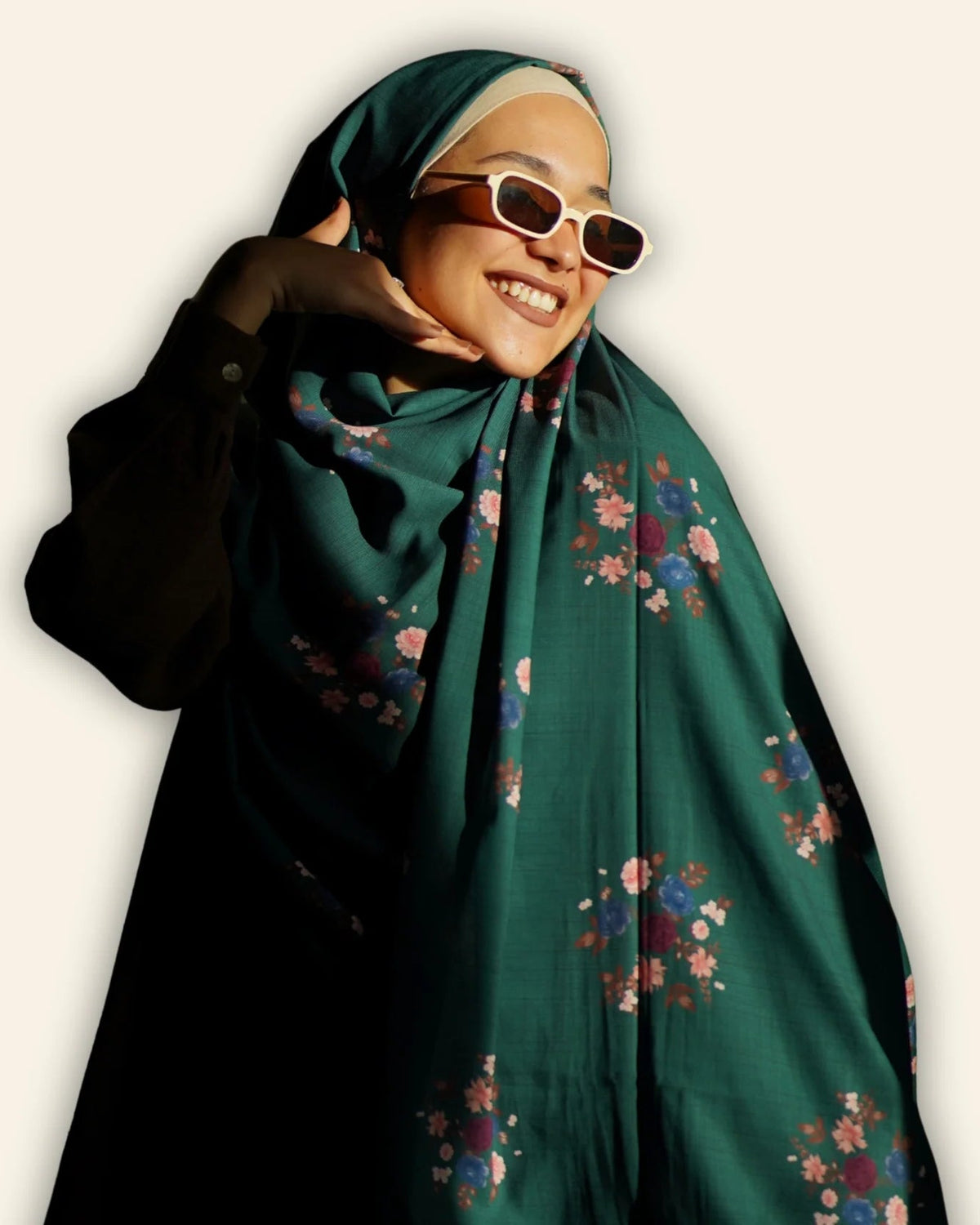 A woman wearing a dark green hijab with a colorful floral pattern, styled with white-framed sunglasses and a black long-sleeve top against a plain off-white background.