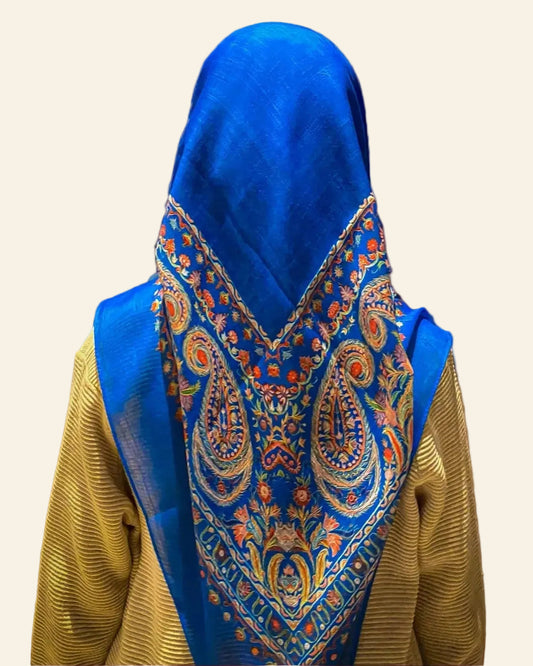 Person wearing a blue and patterned shawl in front of an ornate wall tapestry.