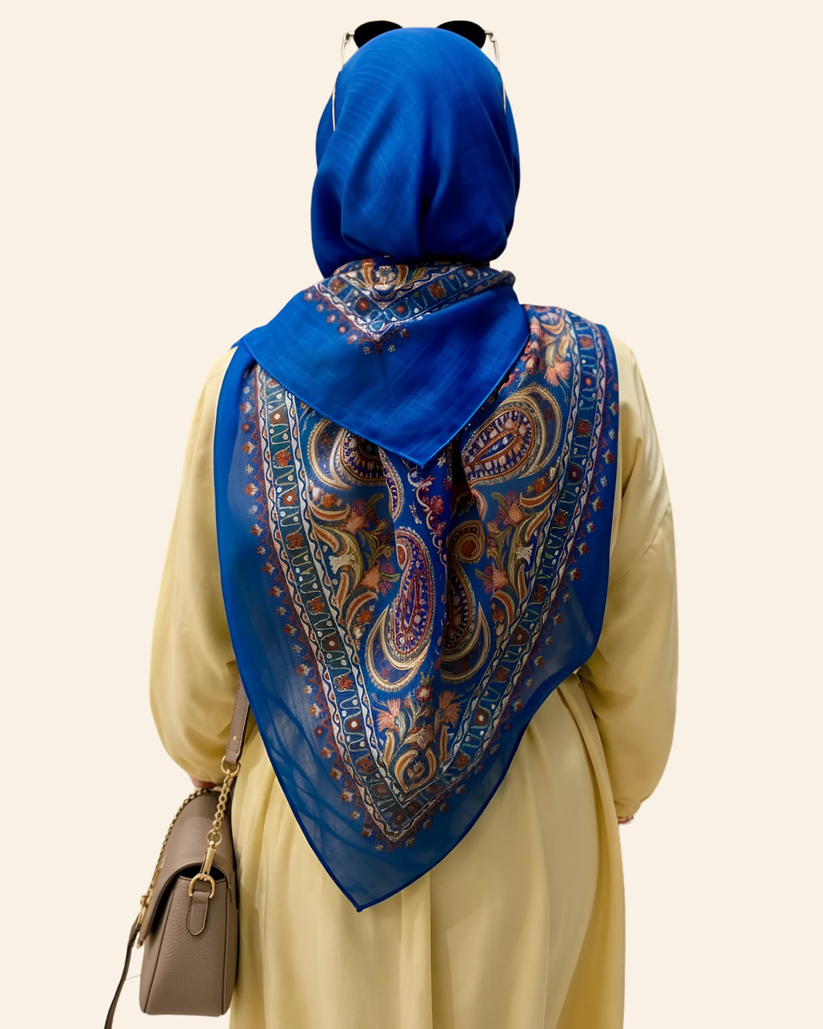 A woman shown from behind wearing a bright blue hijab and a matching paisley scarf draped over her shoulders, paired with a cream-colored dress and a small taupe handbag, against a solid light beige background.