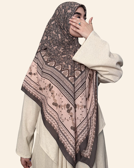 A woman wearing a beige long-sleeve dress and a patterned hijab. The hijab features a dark brown base with a dusty pink lower section and intricate geometric and floral motifs.