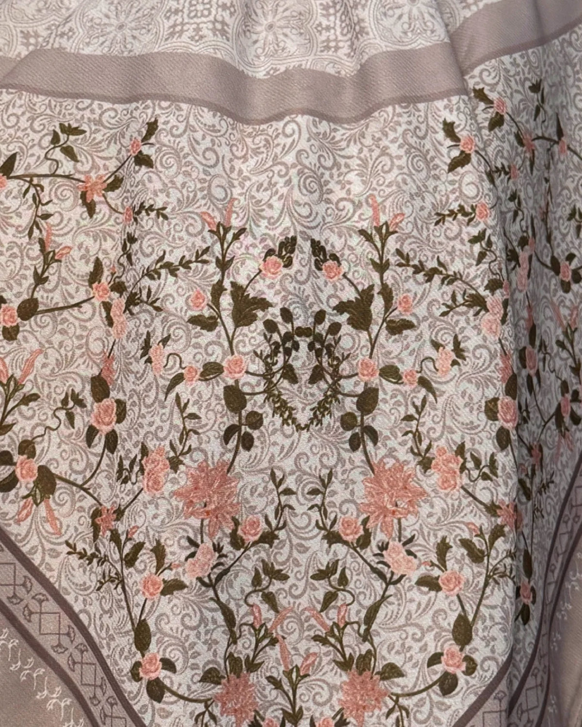 A close-up of an embroidered fabric featuring a detailed floral pattern with pink flowers and dark green leaves on a light beige background, finished with a decorative border.