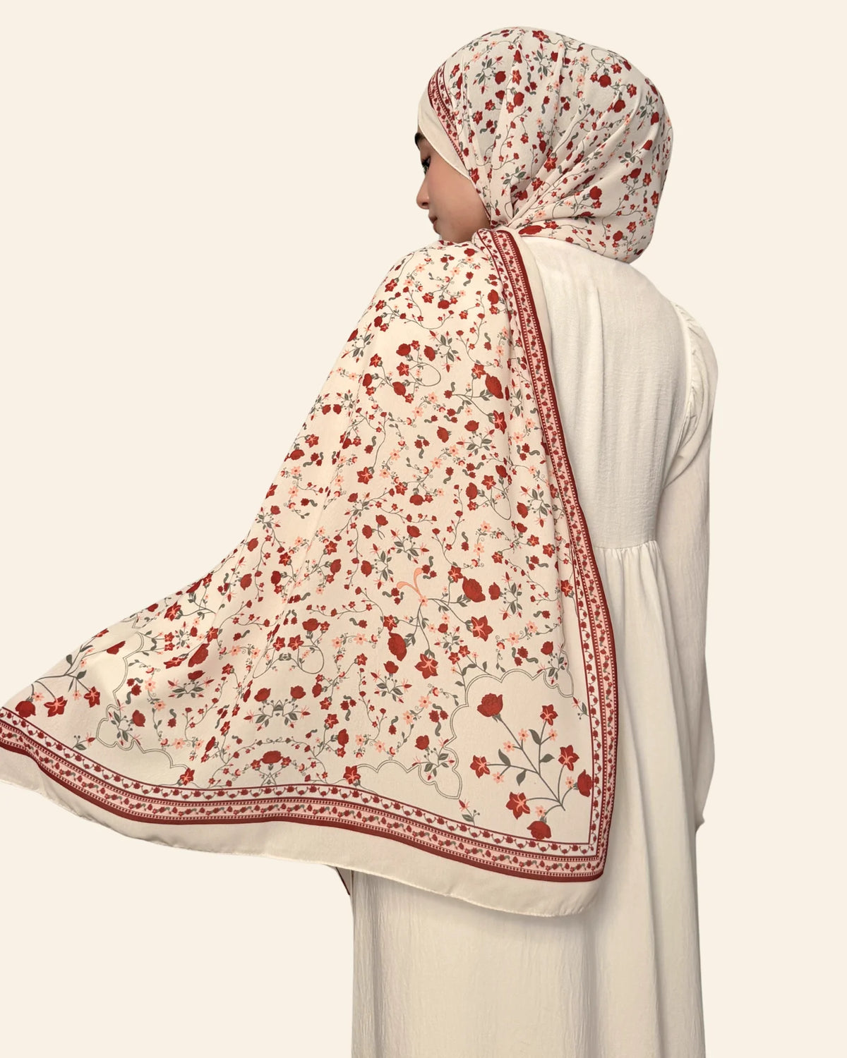 A woman shown from the back wearing a white dress and a cream hijab with a red floral pattern and a decorative red border, set against a solid light beige background.