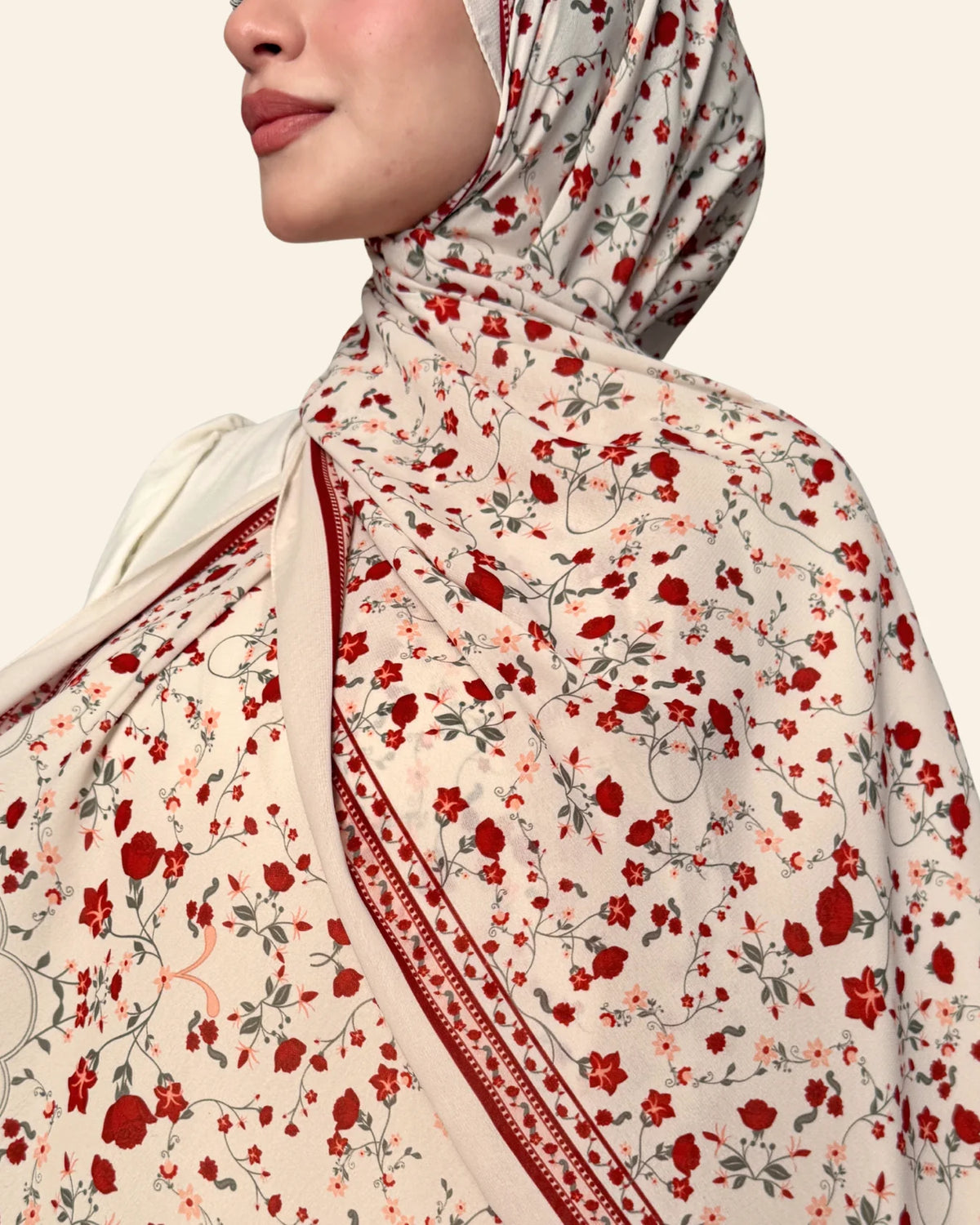 A close-up of a model wearing a beige hijab with a red and green floral print and a decorative red border.