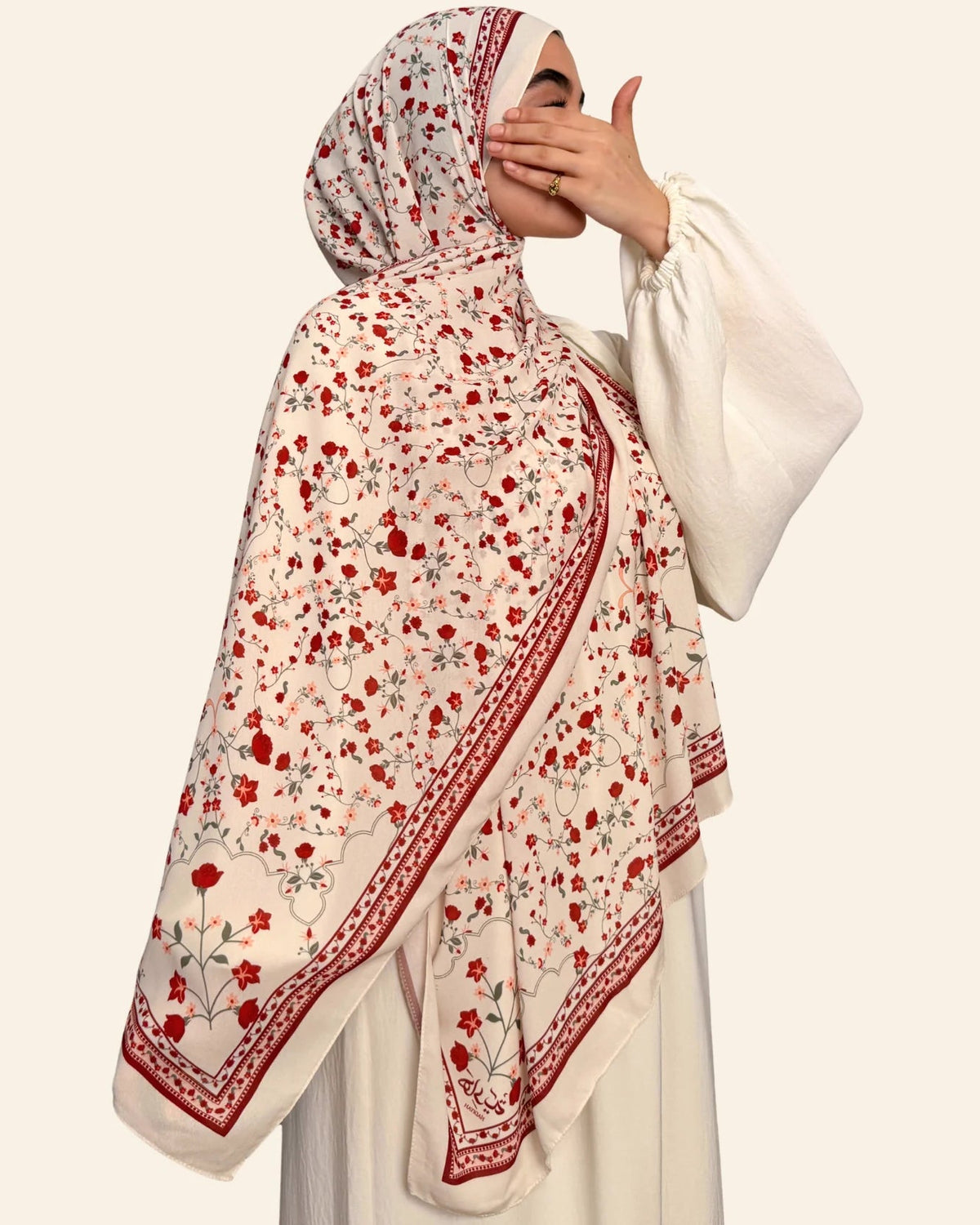 A woman wearing a cream hijab with a red and gray floral print and embroidered border, paired with a white long-sleeve dress against a solid light beige background.