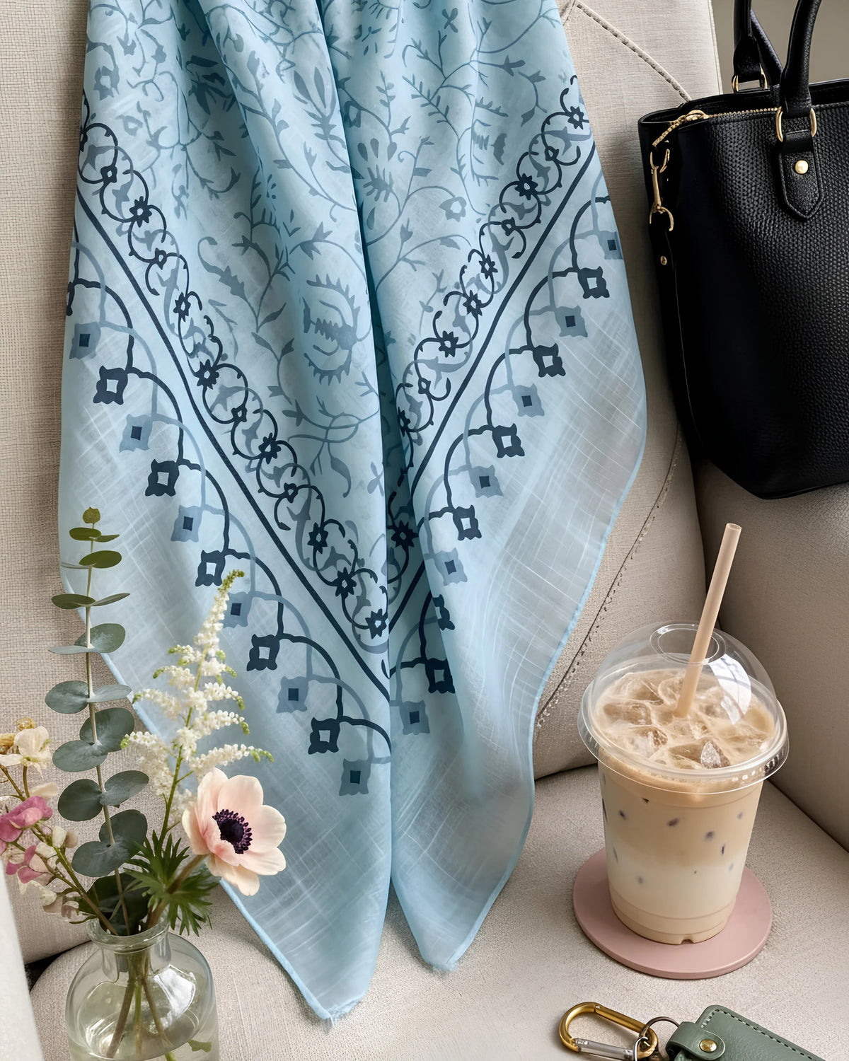 A light blue patterned scarf draped over a beige sofa next to a black handbag, an iced coffee, and a small vase of flowers.