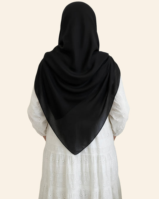 A person shown from the back wearing a black hijab draped over a white eyelet dress, against a solid light beige background.
