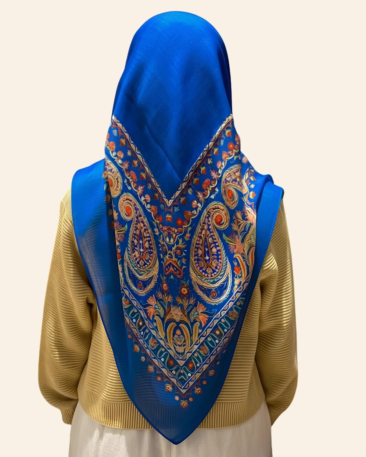 A person shown from the back wearing a bright blue headscarf with an ornate paisley and floral pattern in gold, orange, and white, layered over a gold ribbed sweater.