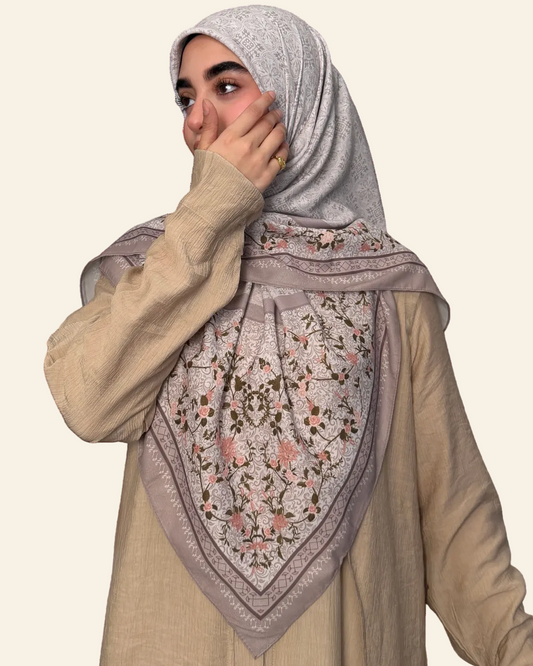 A woman wearing a light beige ribbed long-sleeve top and a light gray patterned hijab with a large matching square scarf featuring pink and brown floral embroidery, posed against a solid white background.