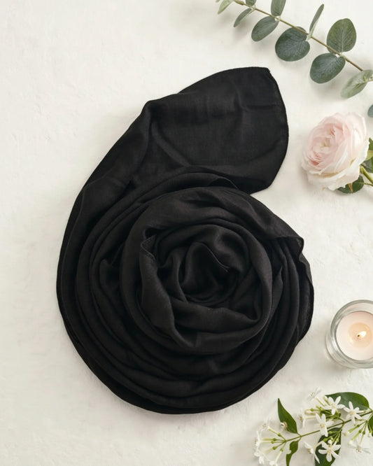 A black lightweight scarf or shawl is arranged in a loose spiral on a light textured surface, styled with green eucalyptus leaves, a pale pink rose, and a small lit candle.